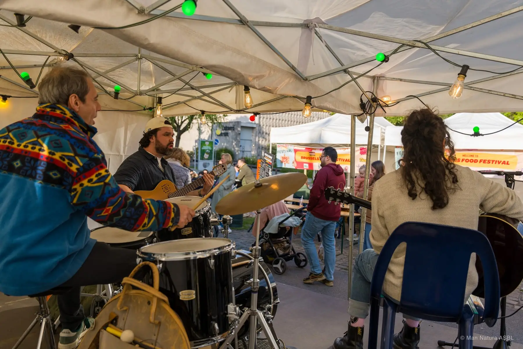 Un groupe joue en acoustique sous une tente du festival, batteur et guitaristes entourés de guirlandes lumineuses