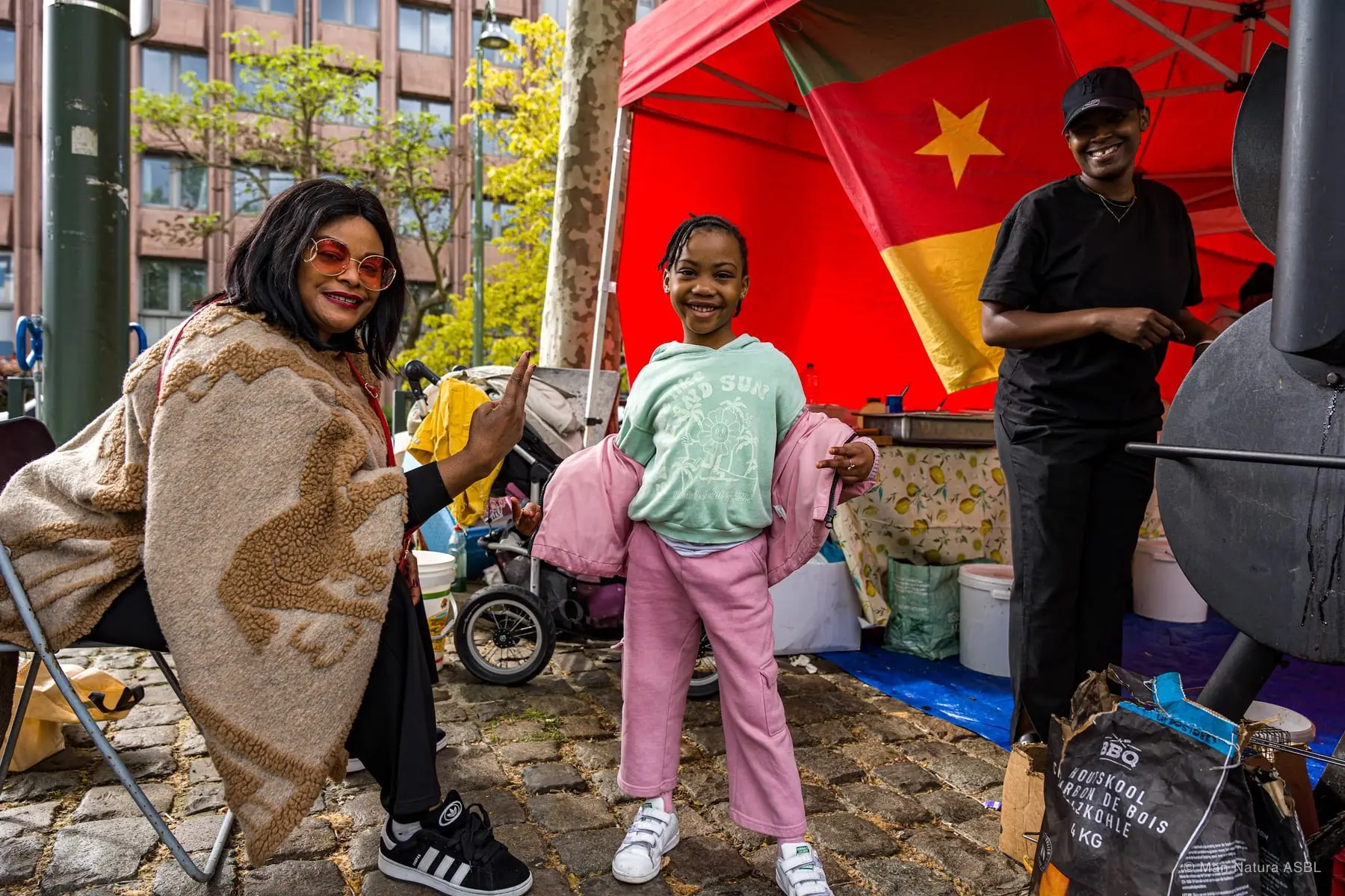 Une mère et sa fille devant le stand camerounais, drapeau rouge et étoile jaune au-dessus du grill