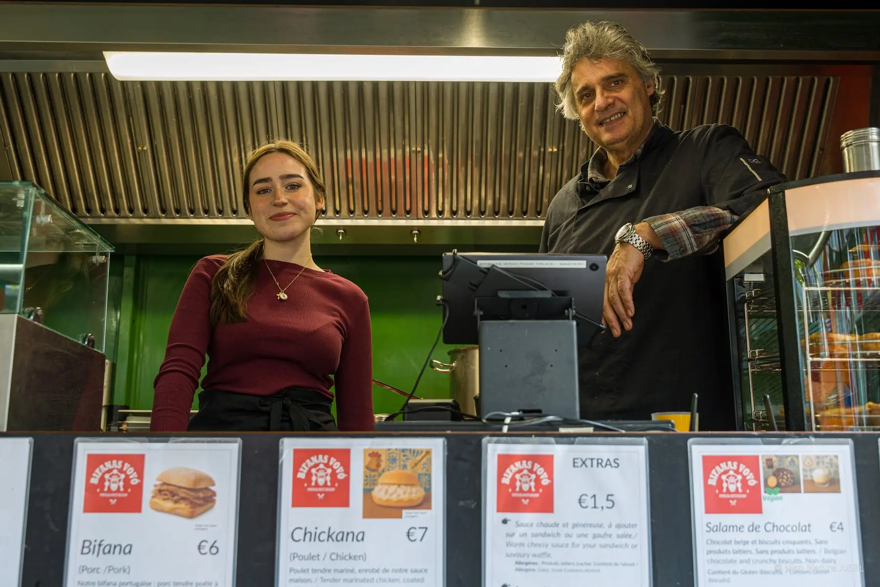 Deux vendeurs derrière leur comptoir portugais, menu bifana et chickana affiché devant
