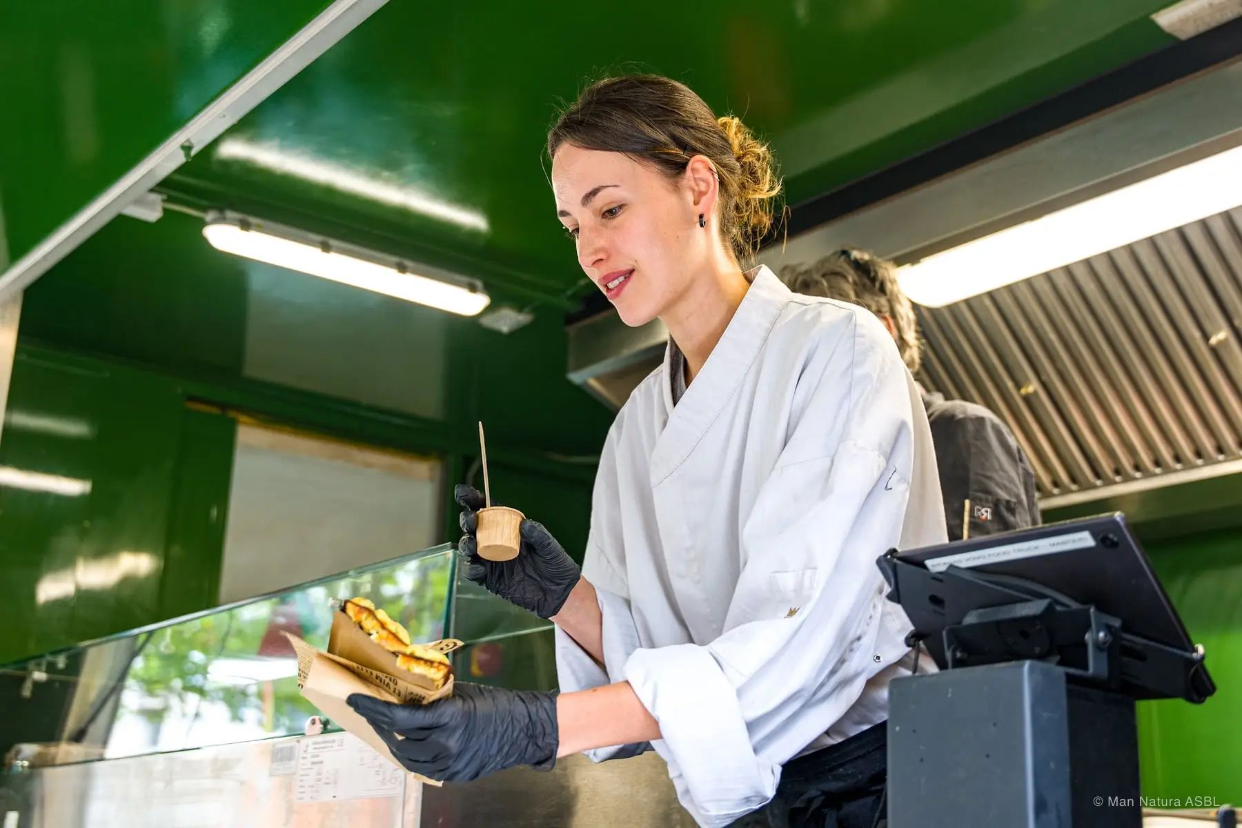 Une vendeuse en veste blanche prépare un sandwich derrière la vitre du camion