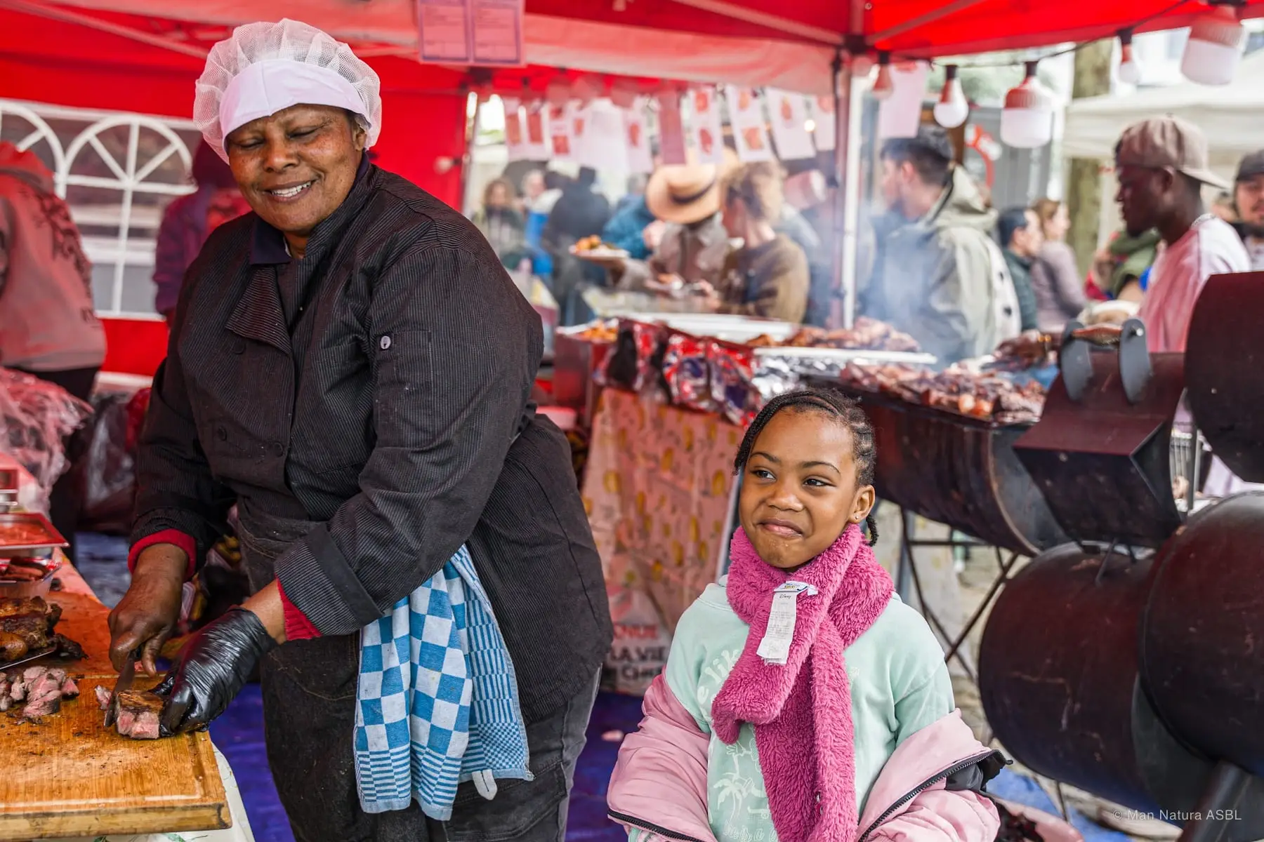Une cuisinière sourit devant son grill, une fillette en écharpe rose à ses côtés
