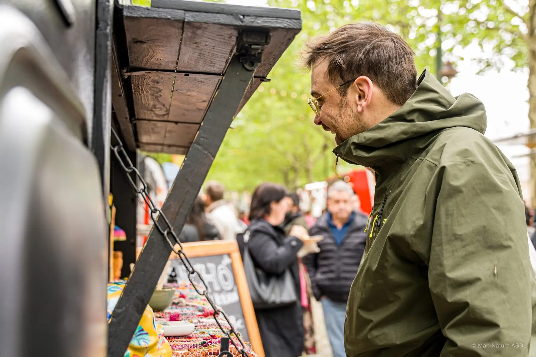 Un visiteur consulte l'ardoise d'un stand, l'allée animée derrière lui