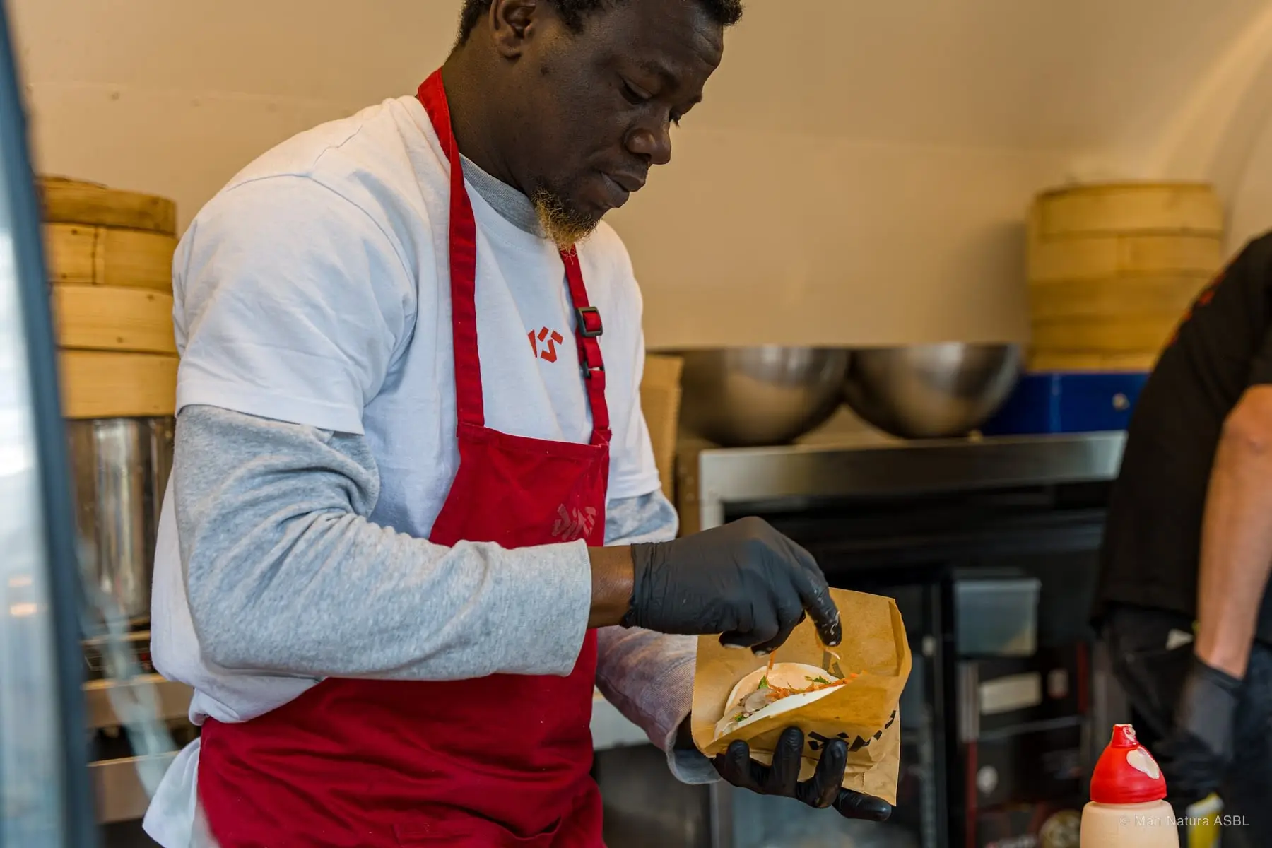 Un cuisinier en tablier rouge garnit un sandwich devant ses paniers vapeur