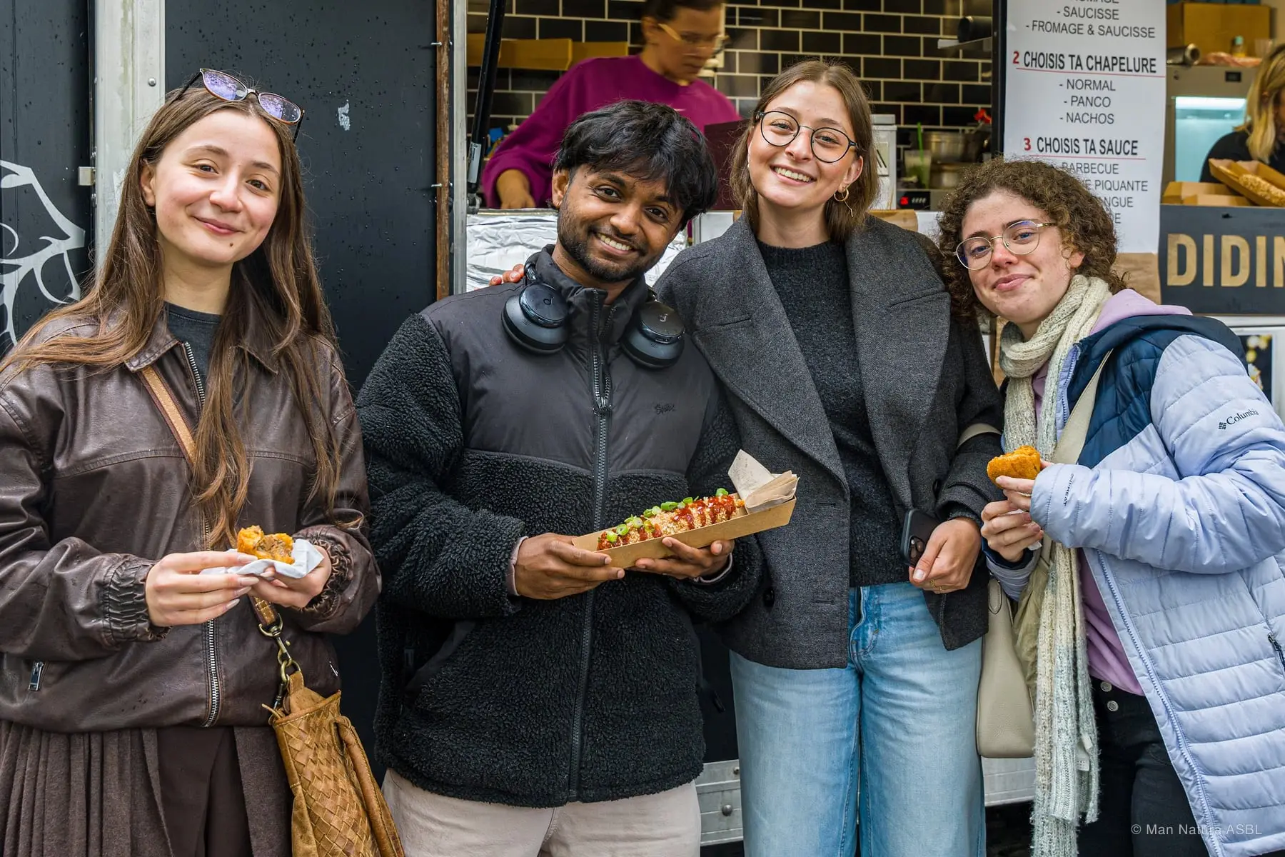 Quatre amis présentent leurs plats devant un stand de saucisses